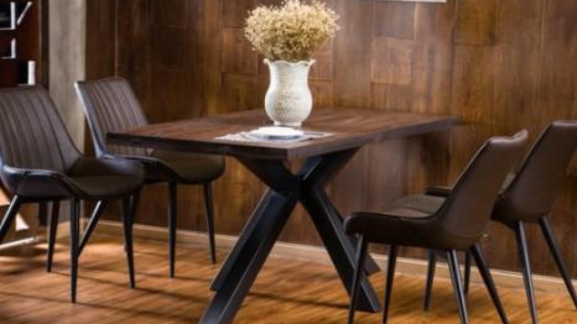 Large wooden table with flower vase on it surrounded by wooden chairs. The room is painted dark brown with wooden flooring.