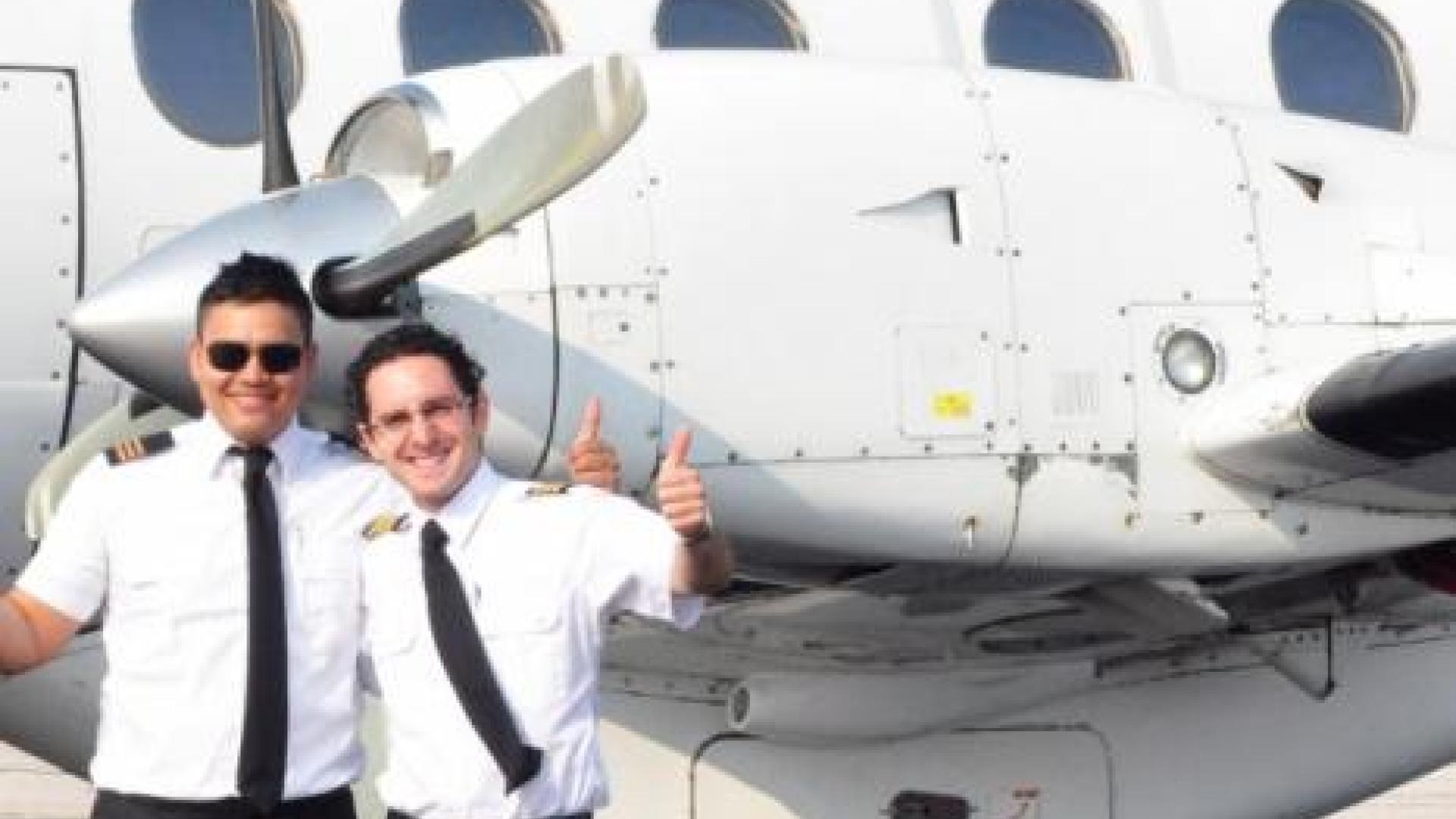 Two airplane pilots smiling and giving a thumbs up. There is a propeller airplane behind the airplane pilots.