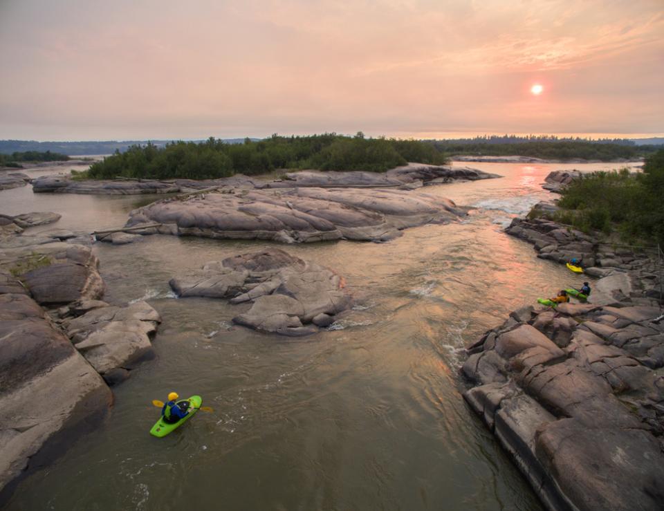 Aerial view of person kayaking through rocky shote. The sun is setting in the background giving the sky an orange glow.