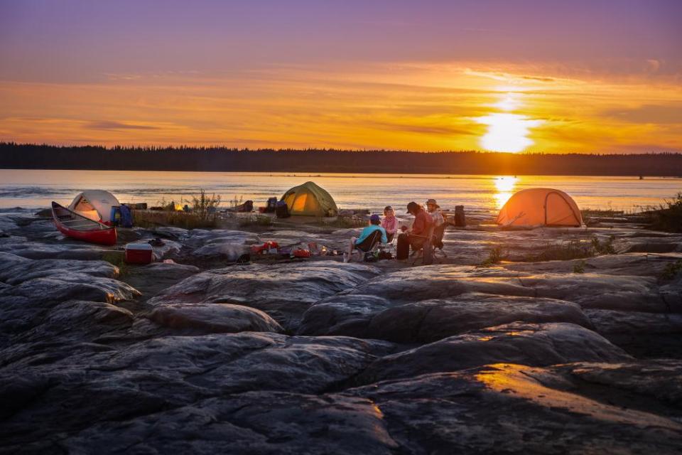 Camping tents set up on rocky shore. The sun is setting in the background as the campers are gathered in a group.