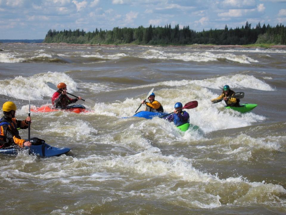 Group of kayakers navigating through river rapids. The kayakers are dressed in and wearing bright-coloured gear.