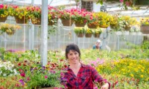 Person smiling as they stand behind elevated flower beds inside a greenhouse. There are flowers everywhere around the greenhouse.