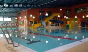 Interior of indoor pool facility. There is a large yellow water slide on the back end of the pool and some swimming lanes on the near side of the pool.
