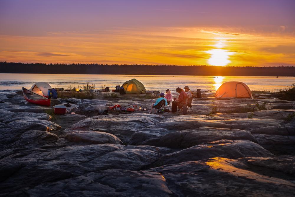 tents by the waterline and a group sitting in camping chairs while the sun sets behind them